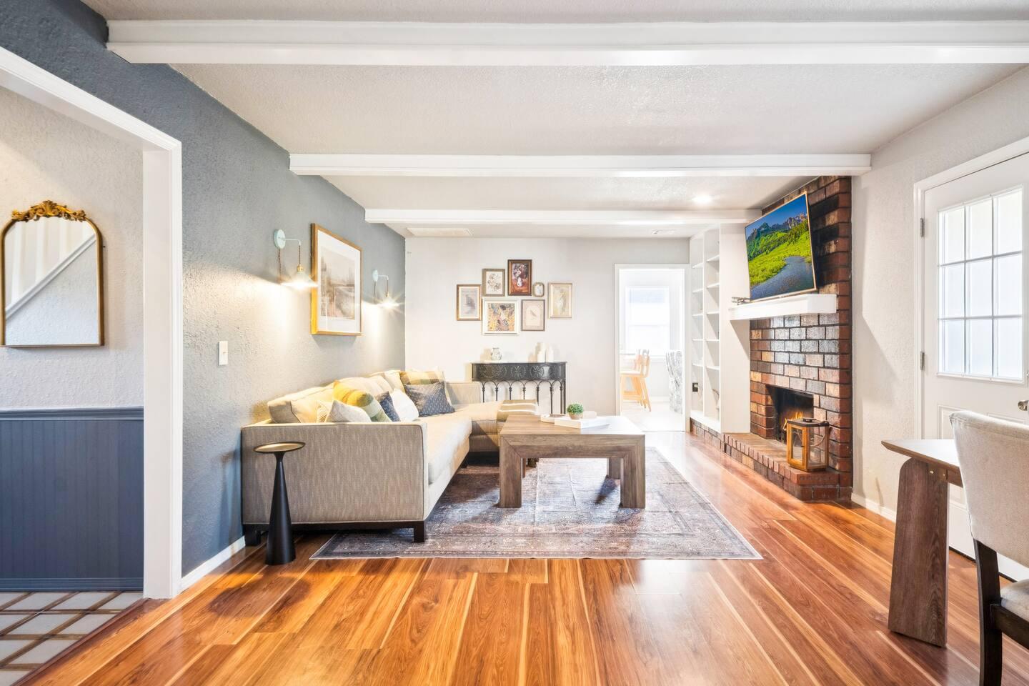 Living room – A bright space with a sectional sofa, wooden coffee table, brick fireplace with a mounted TV, and wooden floors.