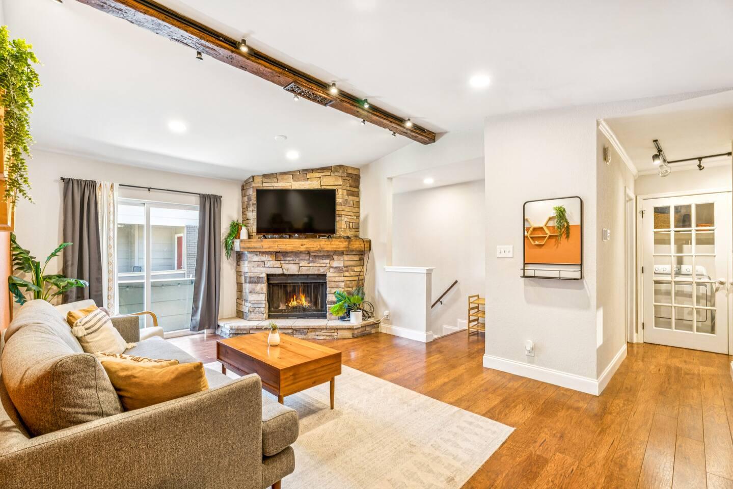 Living Room – Warm and welcoming with a rustic stone fireplace, wood beam ceiling detail, and cozy seating centered around a smart TV. Natural light pours in from the balcony, highlighting the rich hardwood floors and modern farmhouse charm.
