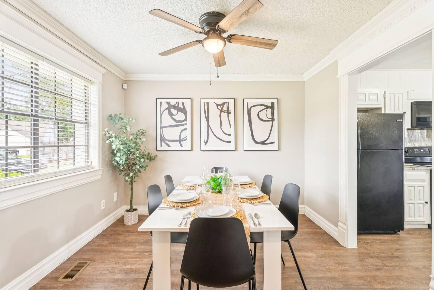 Dining Area – Set for six with modern tableware, sleek black chairs, ceiling fan, abstract wall art, and natural lighting from adjacent kitchen.