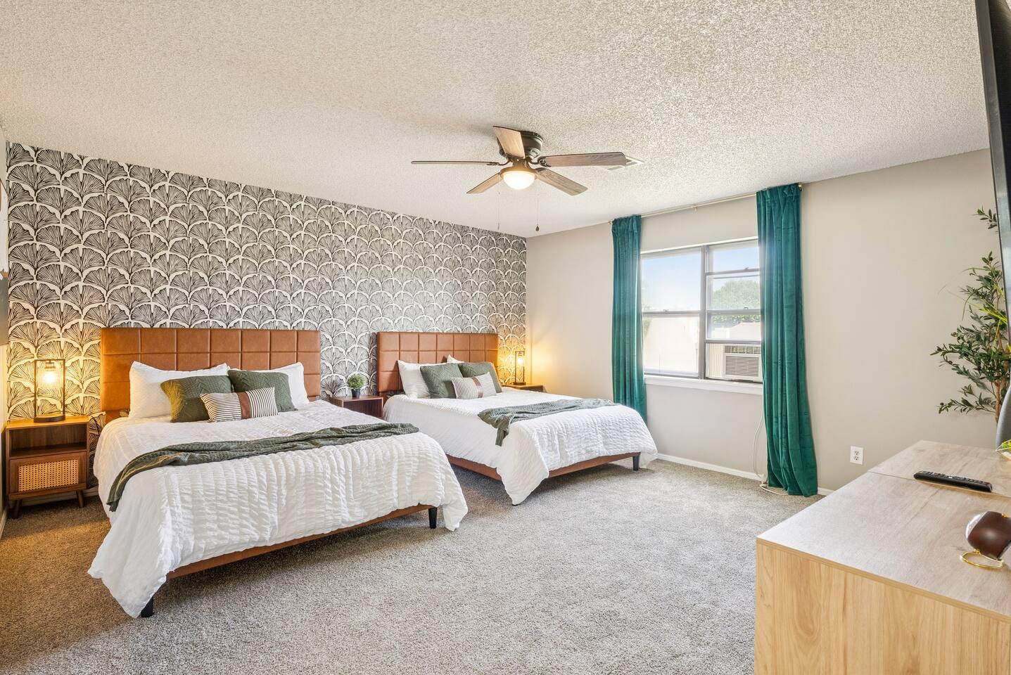 Bedroom – Spacious room with two queen beds, modern wallpaper accent wall, ceiling fan, large window with blackout curtains, and bedside lighting.