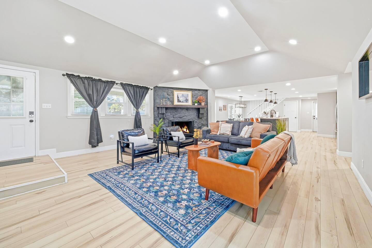 Living Room – Bright open space with light wood floors, a brown leather sofa, dark gray couch, blue patterned rug, and black fireplace. The room has vaulted ceilings, recessed lights, and large windows with dark curtains.