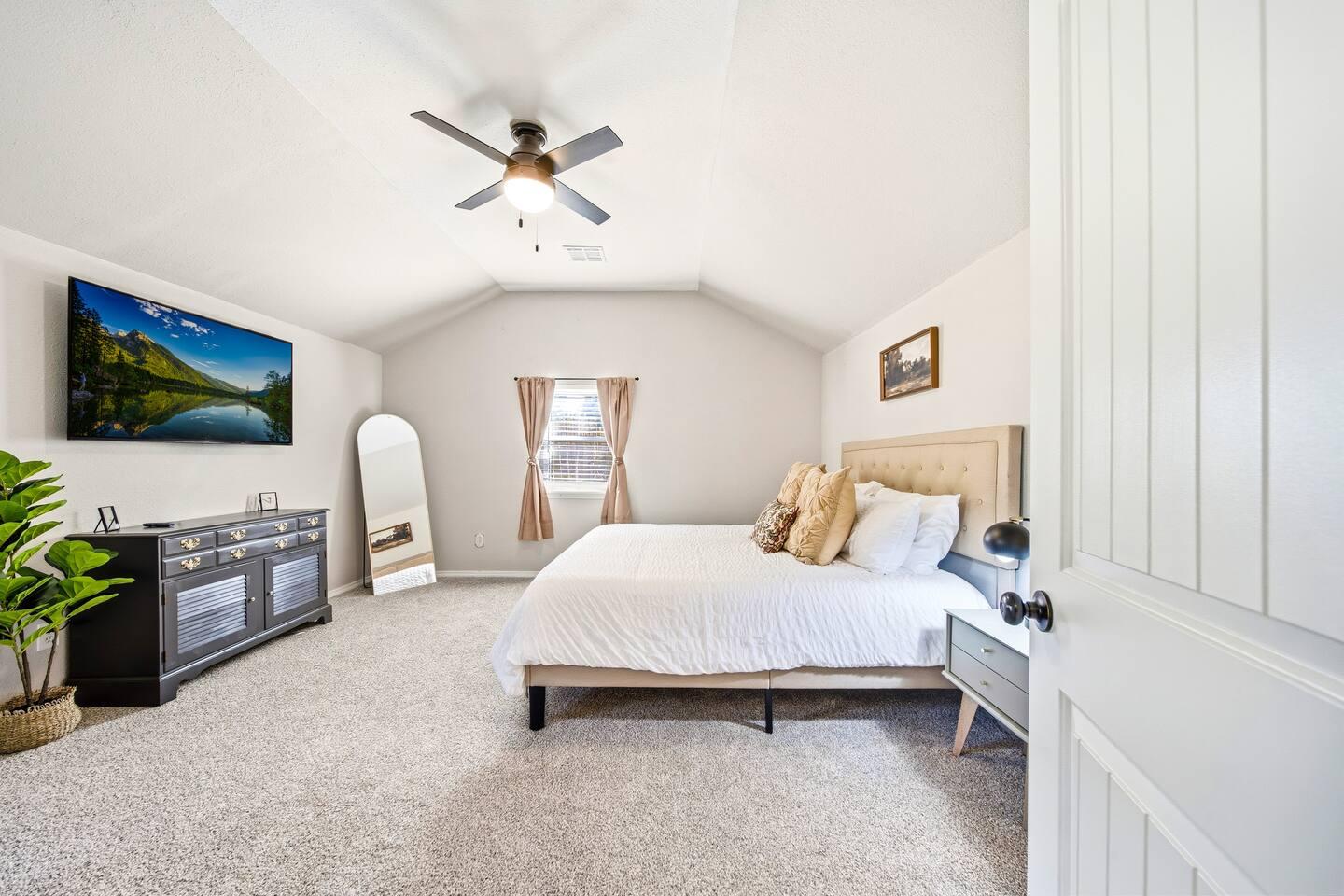 Master Bedroom – Spacious room with a queen bed, soft carpet flooring, and neutral décor. Features a ceiling fan, wall-mounted TV, dresser, and a large mirror beside a window with light curtains.