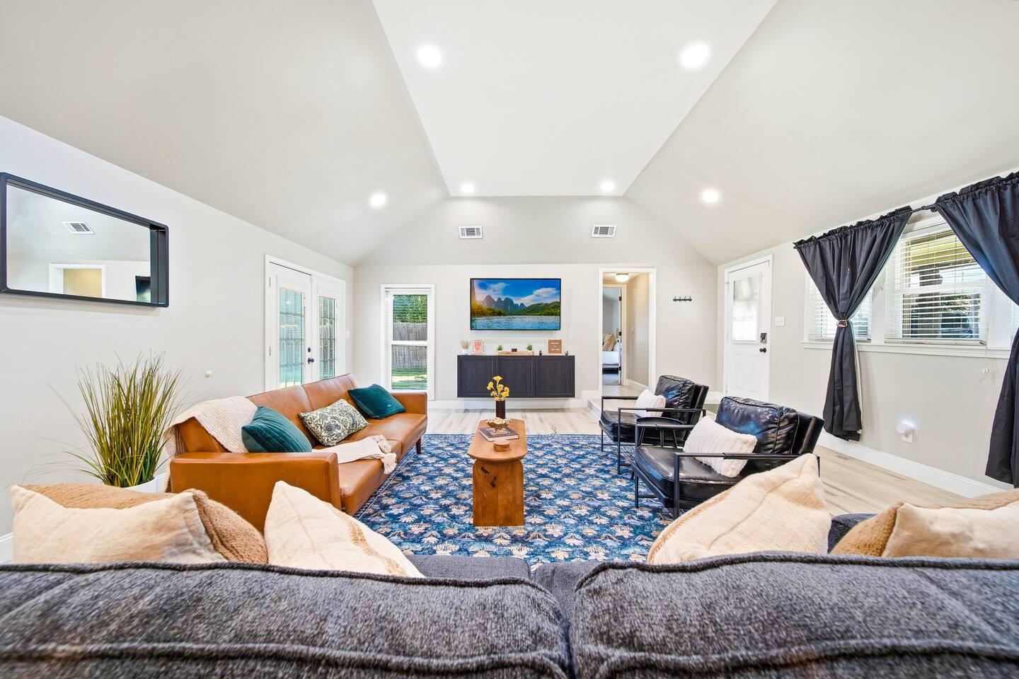 Living Room – Cozy space with a brown leather sofa, two black chairs, and a blue patterned rug. Features a wall-mounted TV, bright windows with dark curtains, and double doors leading to the backyard.