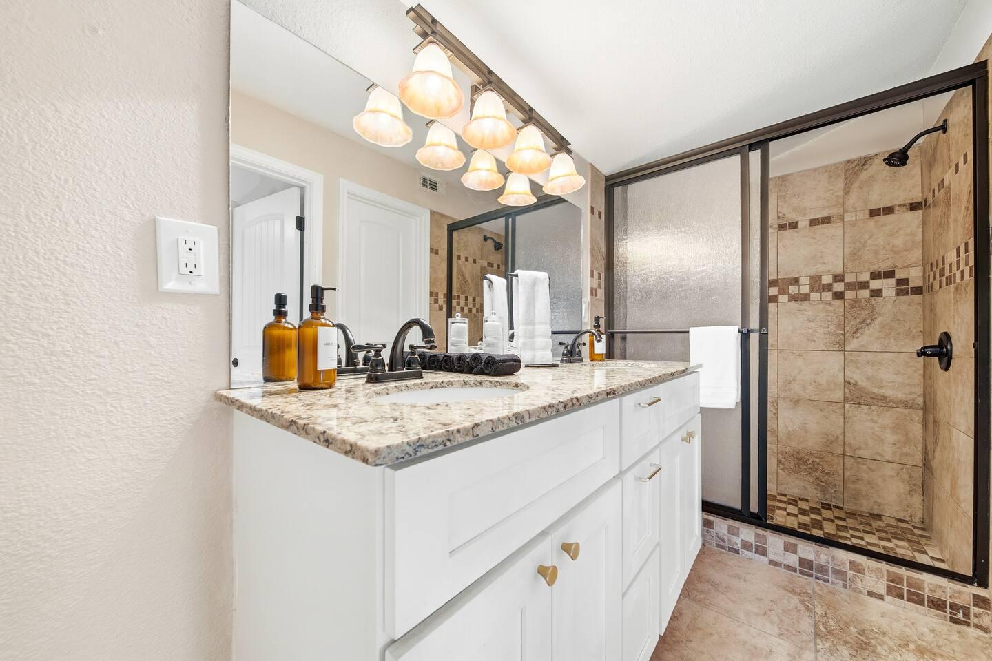 Bathroom – Features a granite countertop vanity with dual sinks, large mirror, and warm lighting. Includes a tiled walk-in shower with glass doors and black fixtures.