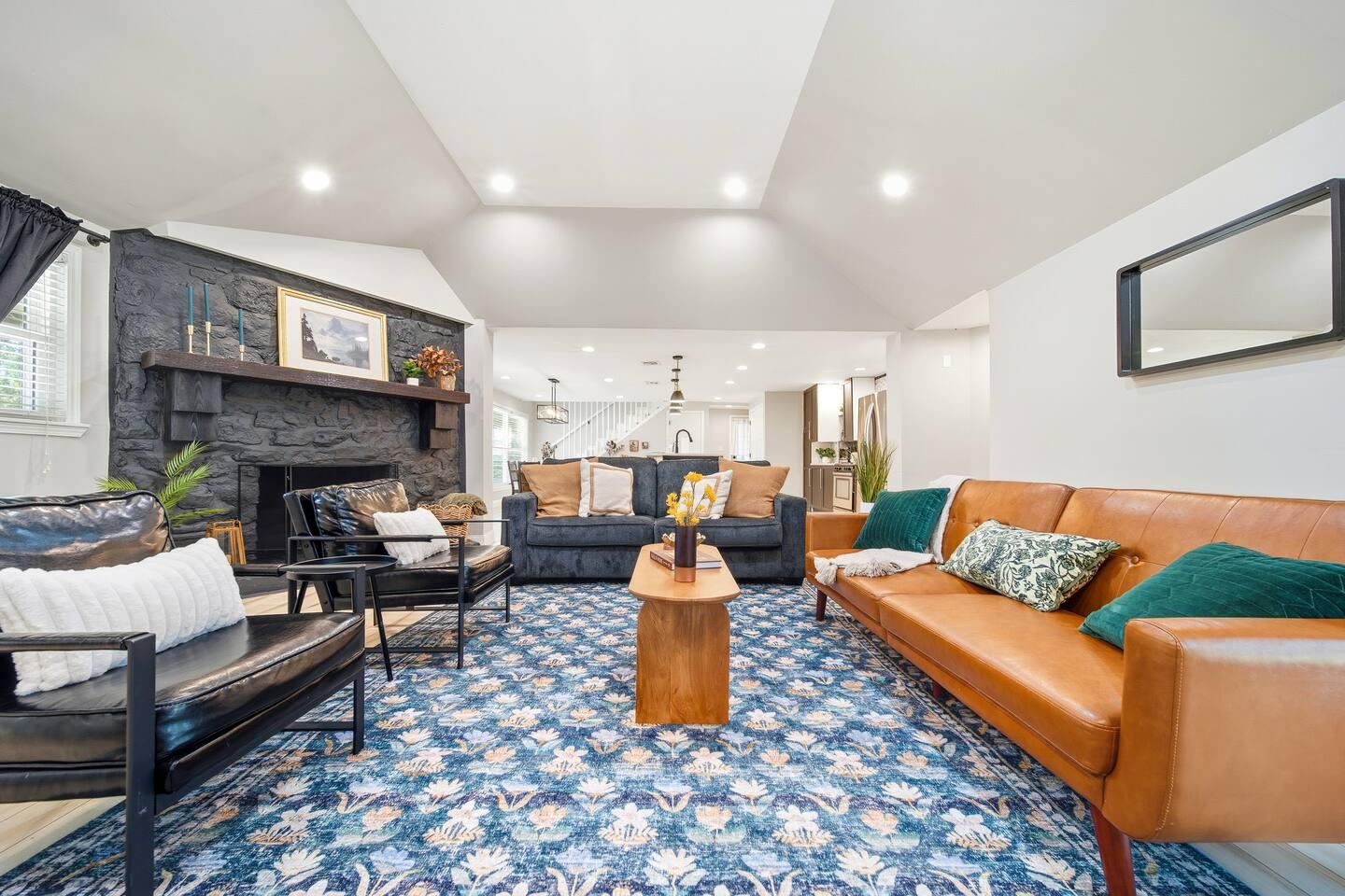 Living Room – Stylish space with a brown leather sofa, dark couch, and two black chairs around a wooden coffee table. Features a blue patterned rug, black stone fireplace, and recessed lighting.