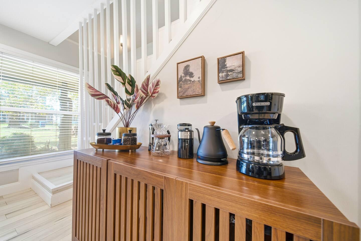 Coffee Corner – Wooden cabinet setup with a drip coffee maker, electric kettle, grinder, and French press. Decorated with plants, jars, and wall art beneath a staircase with natural light from a large window.