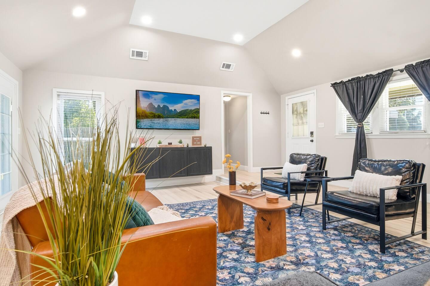 Living Room – Bright space with a brown leather sofa, two black chairs, and a wooden coffee table on a blue patterned rug. Includes a wall-mounted TV, large windows with dark curtains, and double doors leading outside.