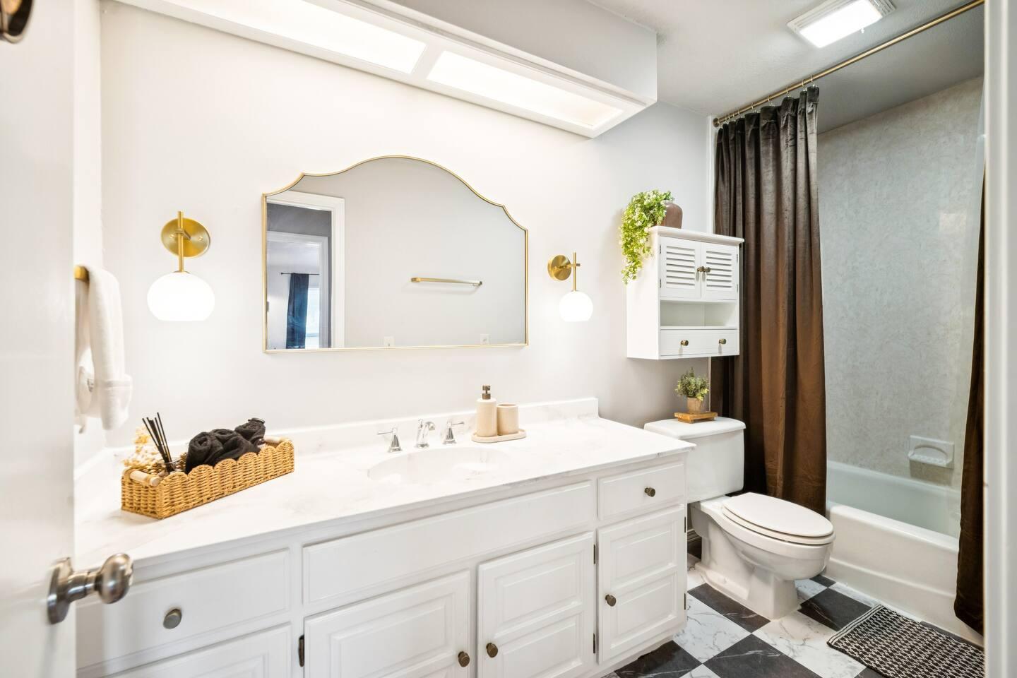 Bathroom – A white bathroom with a large vanity, mirror, toilet, bathtub with a dark curtain, and wall lights.