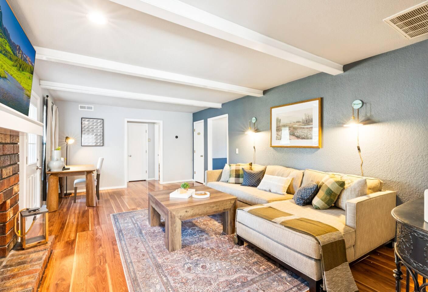 Living Room: A cozy living room with a beige sectional sofa, wooden coffee table, and blue accent wall. A fireplace with a TV above it is visible, and a wooden desk sits near the back wall by the door.