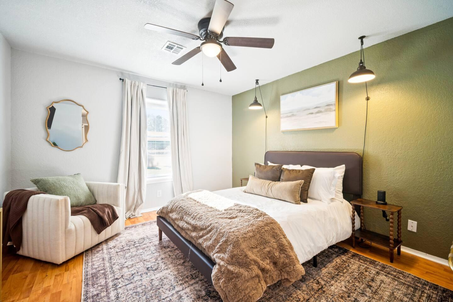 Bedroom – A bed with white bedding, green accent wall, two hanging lights, a ceiling fan, nightstand, armchair, and window with curtains.